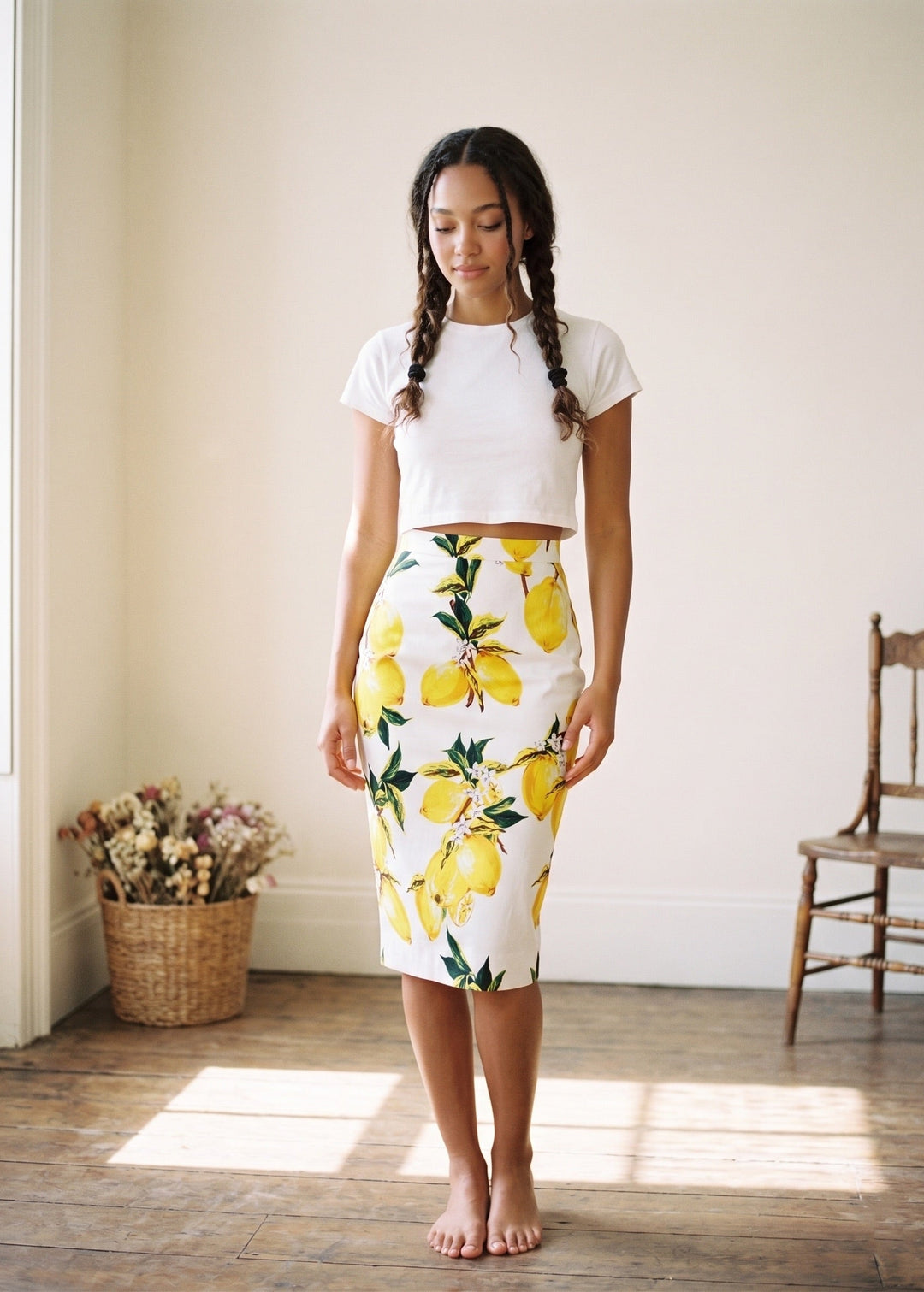Woman wearing a white top and yellow floral skirt standing in a room with wooden floor and white walls.