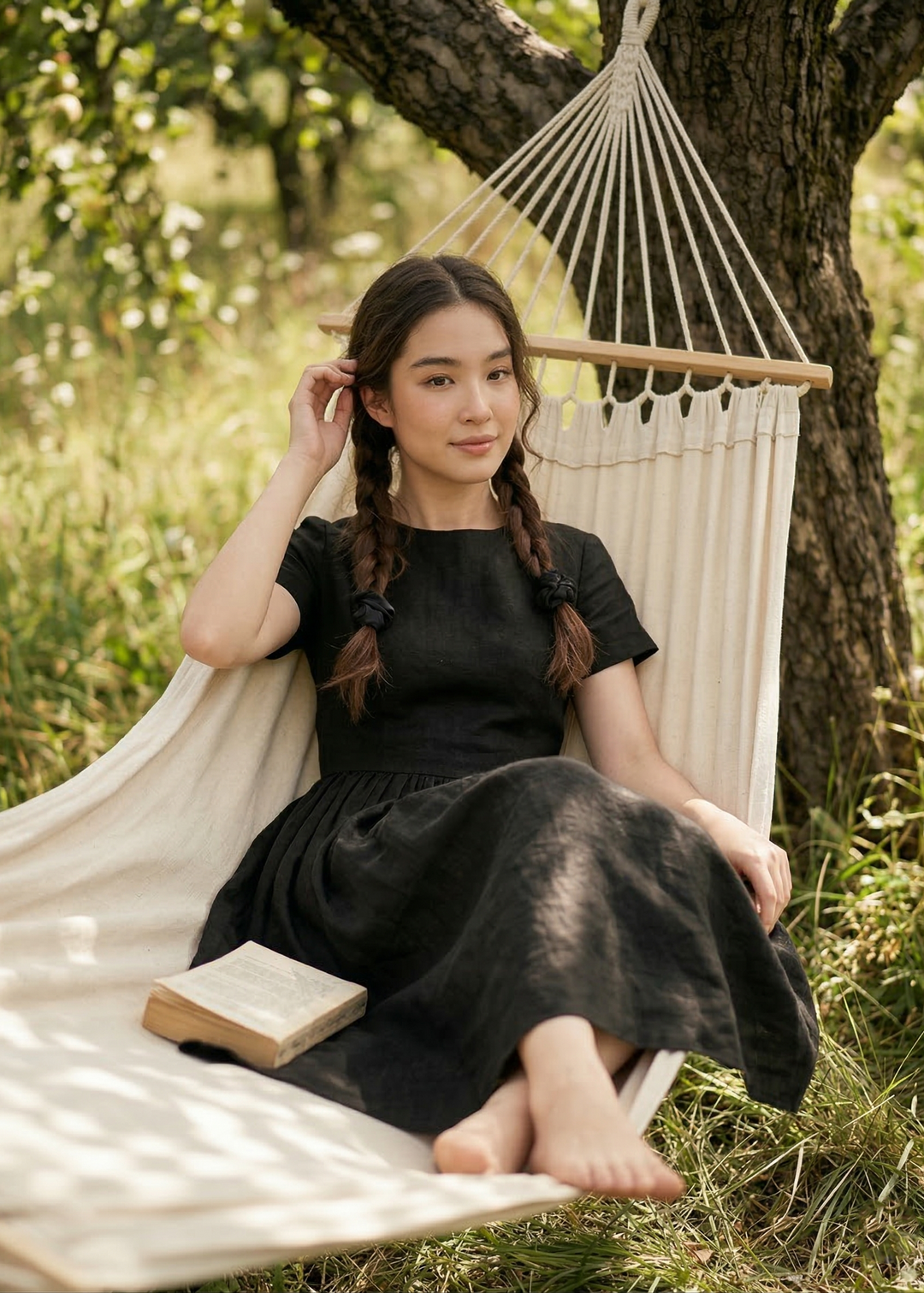 Woman in a black dress sitting in a hammock with a book, surrounded by nature.
