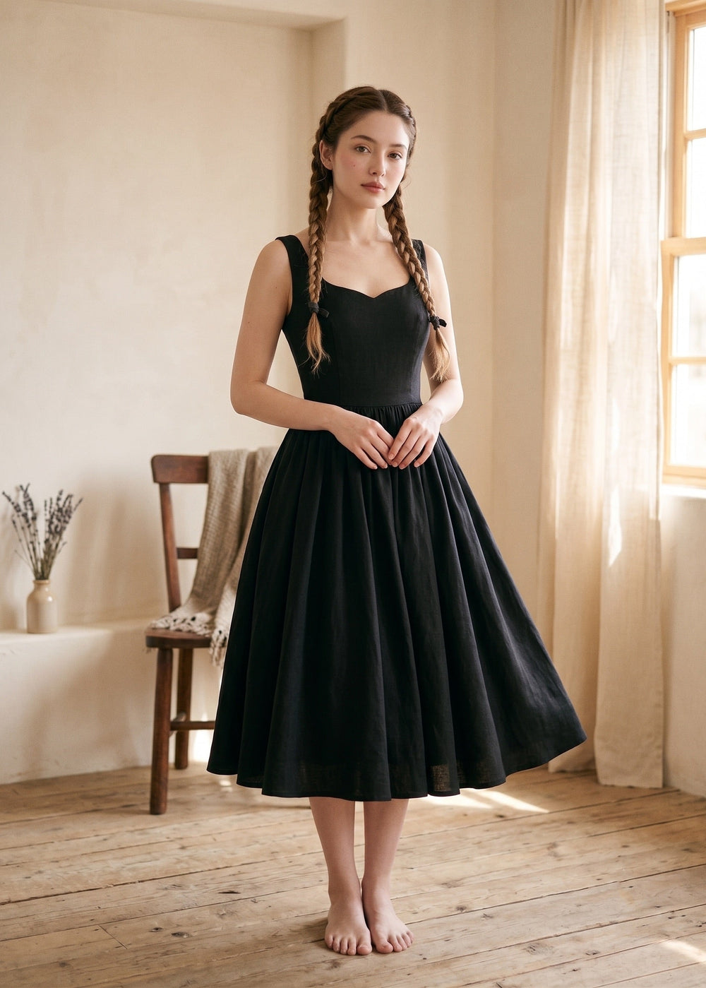 Woman wearing a black dress standing in a softly lit room with wooden floor and window.