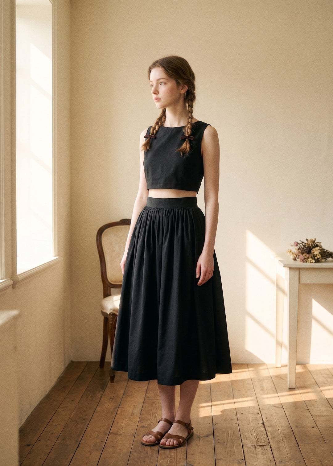 Woman wearing a black dress standing in a softly lit room with wooden floor and window.