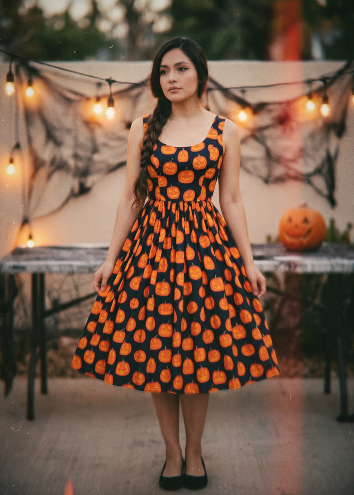 A woman stands outdoors wearing a sleeveless A-line dress with an orange pumpkin print on a black background. The setting has Halloween decorations, including string lights, cobwebs, a table, and a carved pumpkin in the background.