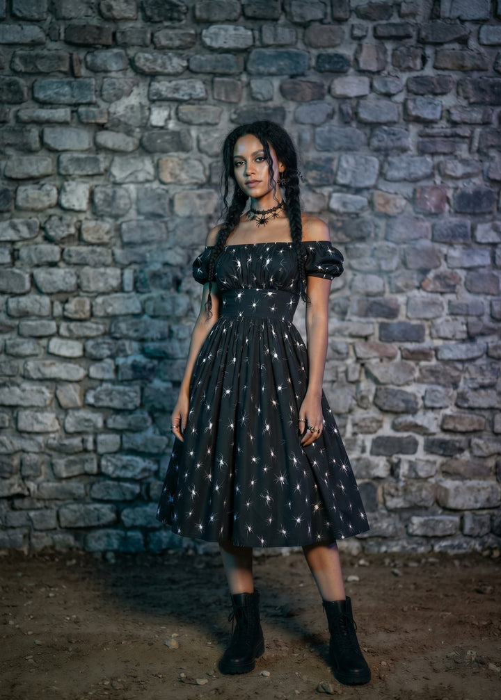 A woman stands in front of a rustic stone wall wearing a black off-the-shoulder midi dress with a white celestial print, paired with black ankle boots.