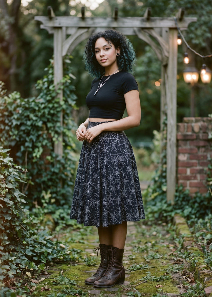 A woman stands outdoors in a garden setting wearing a black short-sleeve crop top and a dark midi skirt with a light geometric pattern, paired with brown lace-up ankle boots.