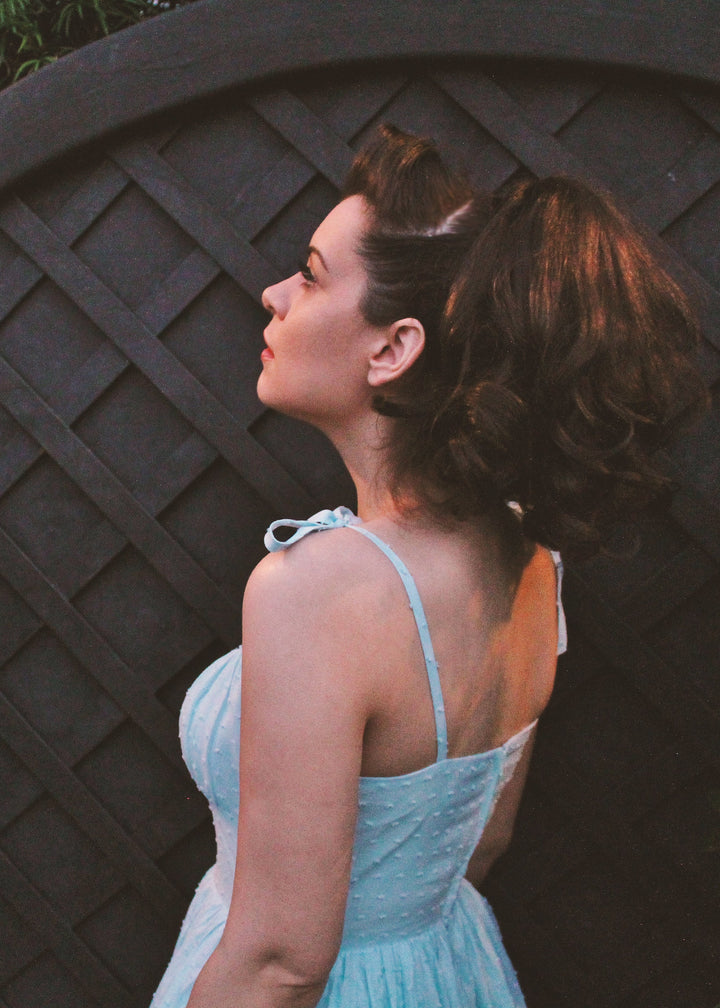 Woman in a light blue dress standing next to a black lattice fence with pink flowers.