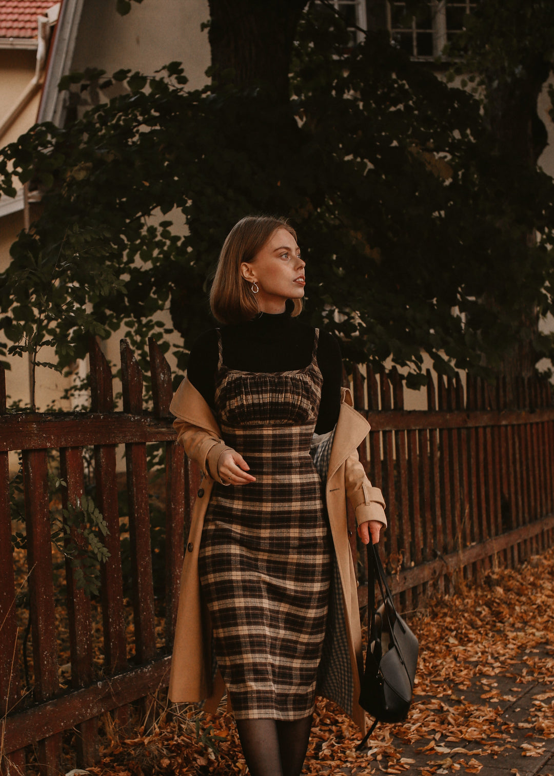 Woman in a plaid dress and beige coat standing near a wooden fence with autumn leaves.