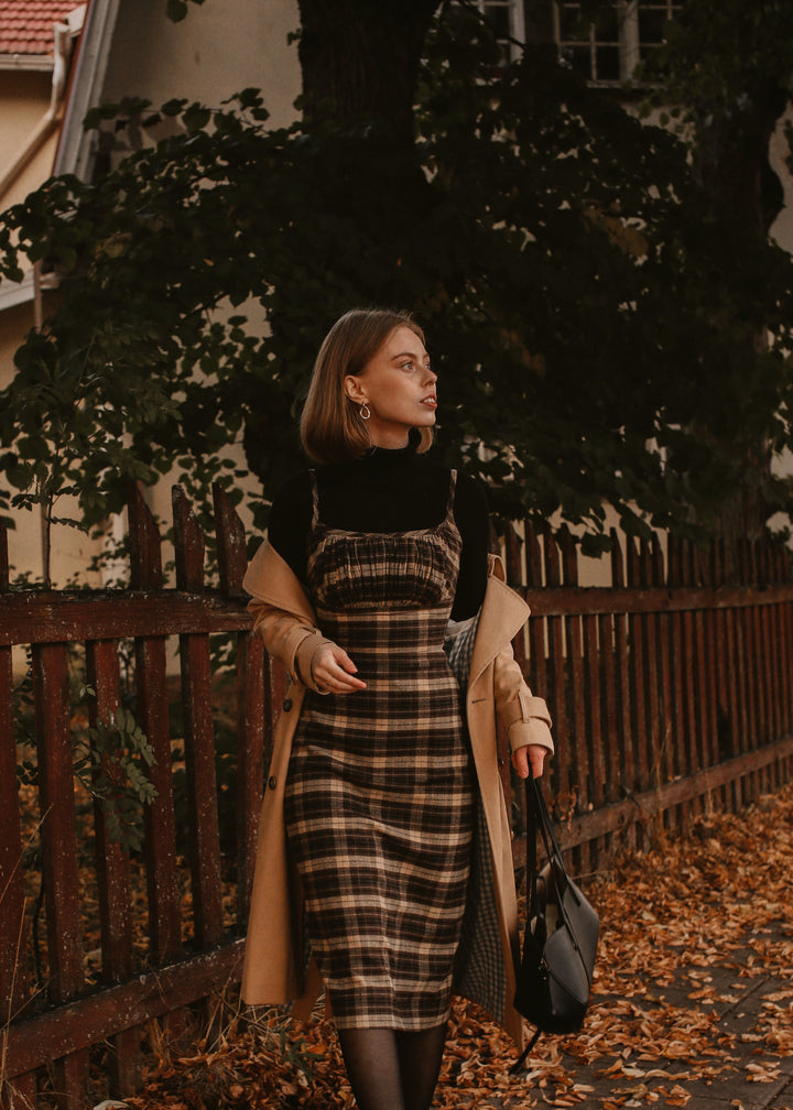 Woman in a plaid dress and beige coat standing near a wooden fence with autumn leaves.