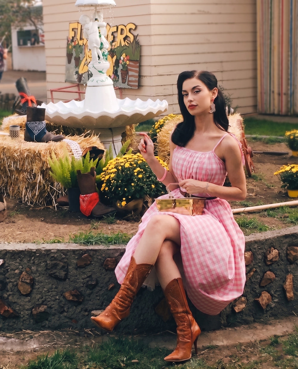 Woman in a pink dress and brown boots sitting outdoors with flowers and a fountain in the background.
