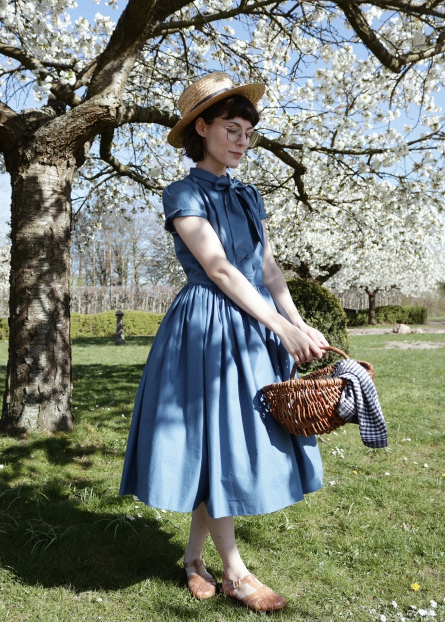 Woman in a blue dress and straw hat holding a basket under a cherry blossom tree.