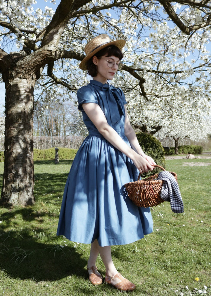 Woman in a blue dress and straw hat holding a basket under a cherry blossom tree.