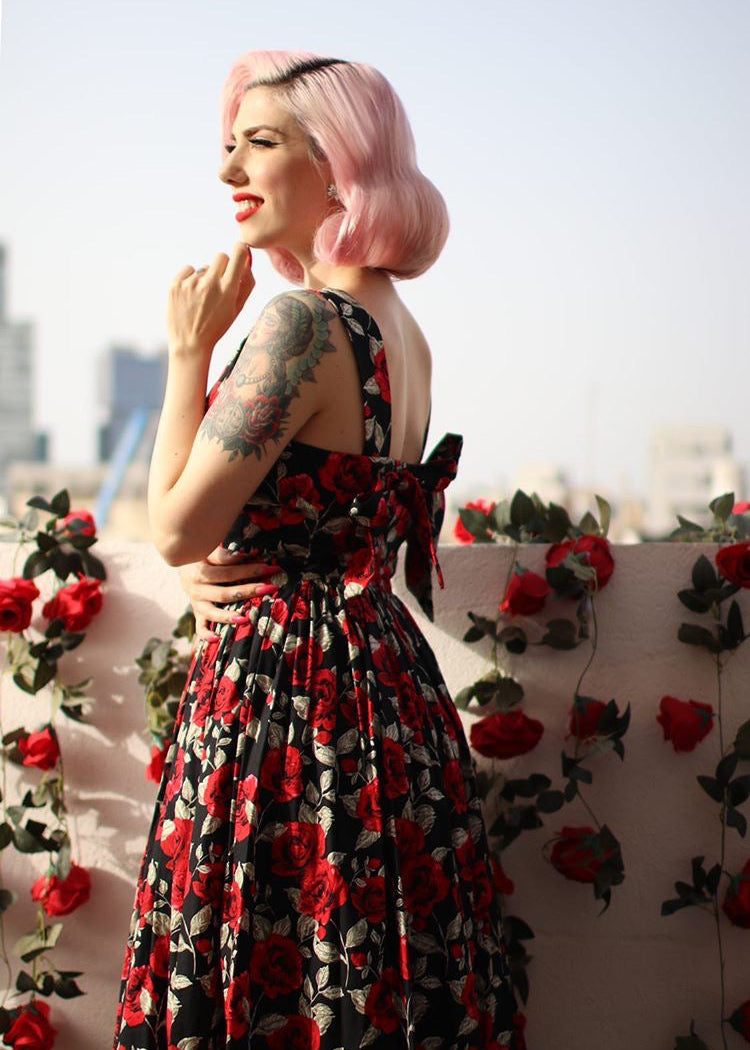 Woman in a floral dress standing against a wall with red flowers and greenery.