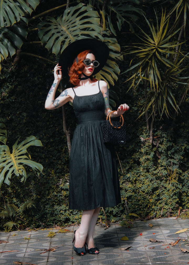 Woman in a black dress and hat standing in front of green foliage