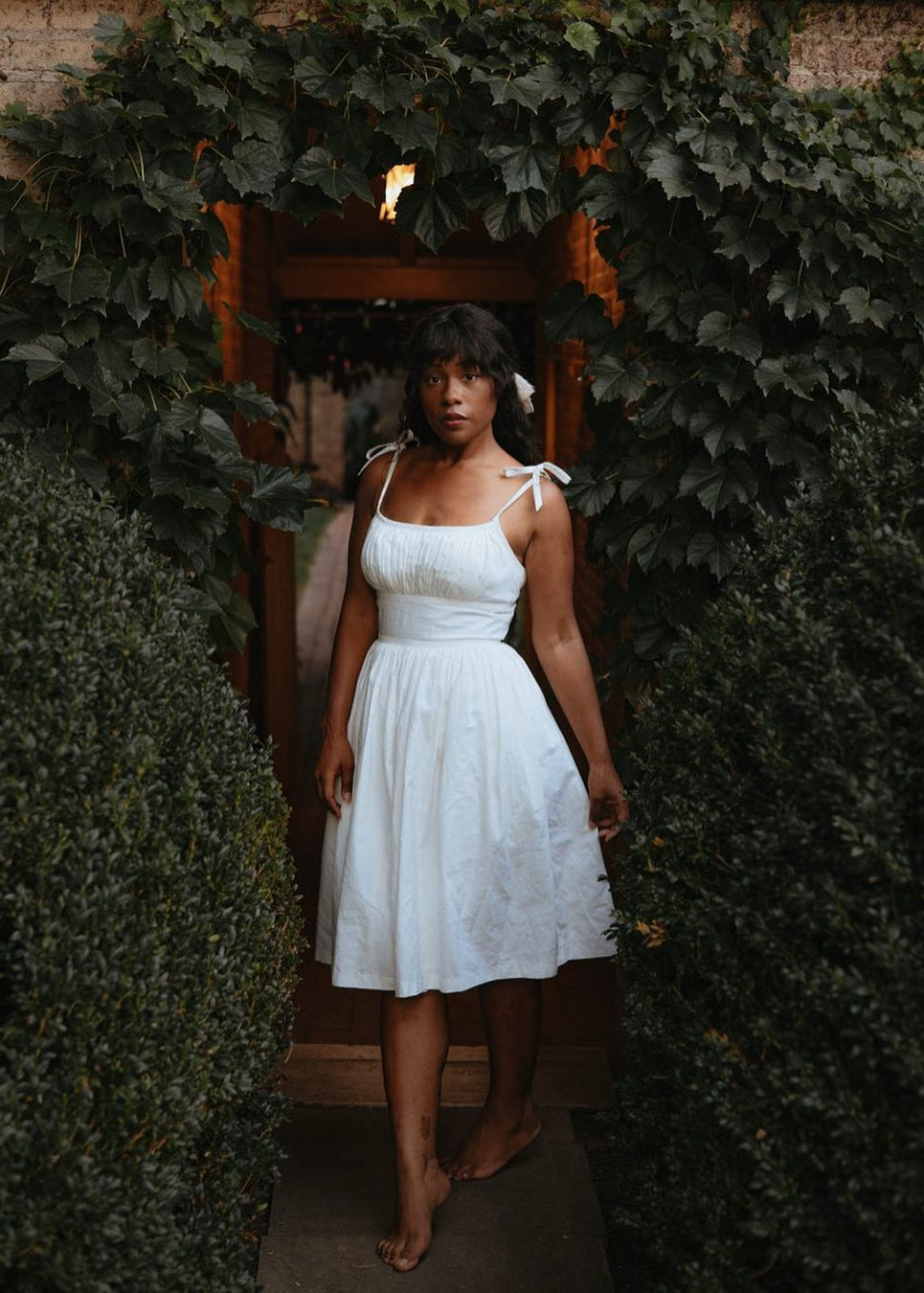 Woman in a white dress standing in front of a wooden door with greenery