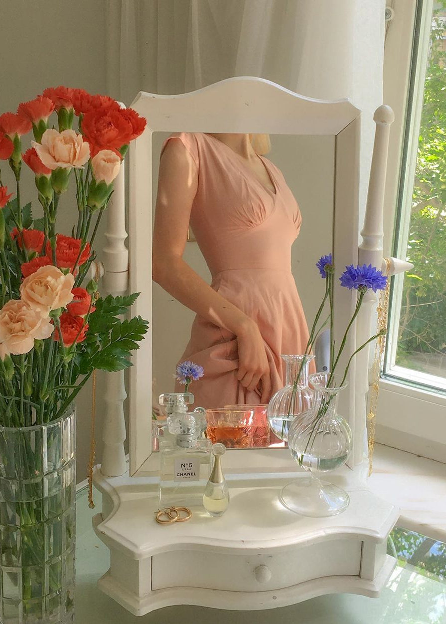 Woman in a pink dress standing in front of a mirror with flowers and perfume on a table.