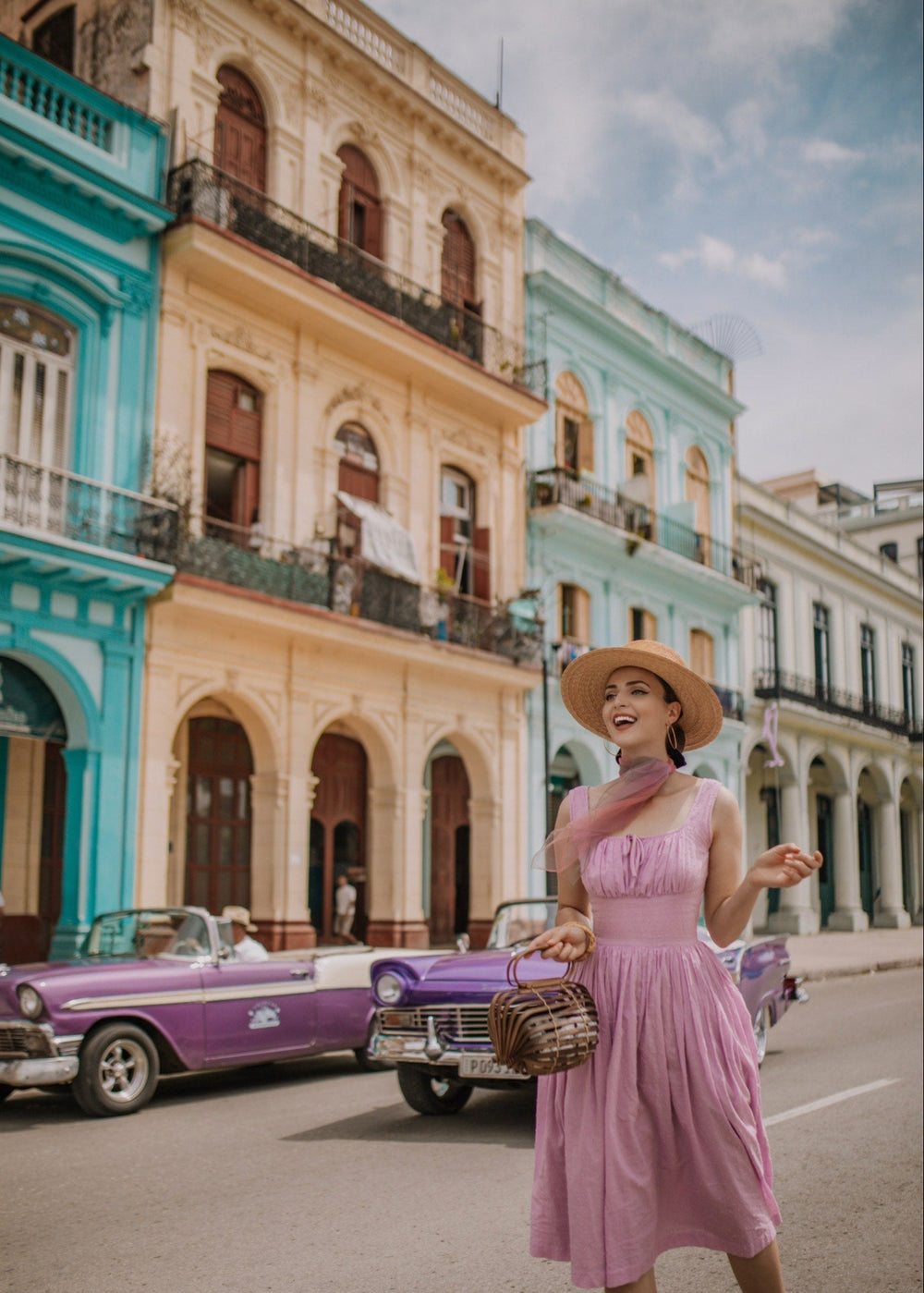 Woman in a pink dress standing on a street with colorful buildings and vintage cars in the background.