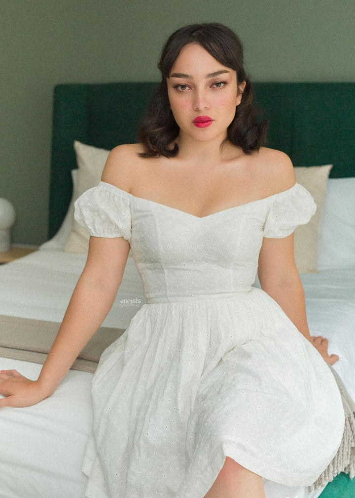 Woman in a white off-shoulder dress sitting on a bed with a green headboard.