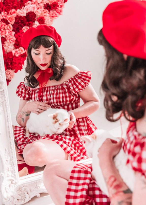 Woman in a red and white checkered dress and beret holding a white cat, sitting in front of a mirror.
