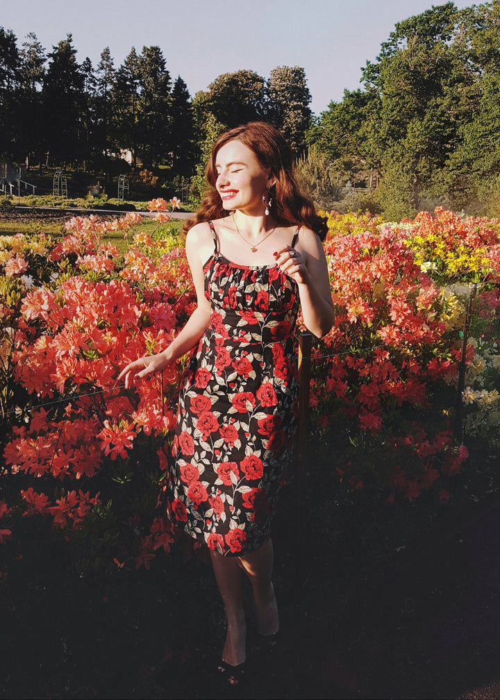 Woman in a floral dress standing in a garden with colorful flowers and trees in the background.
