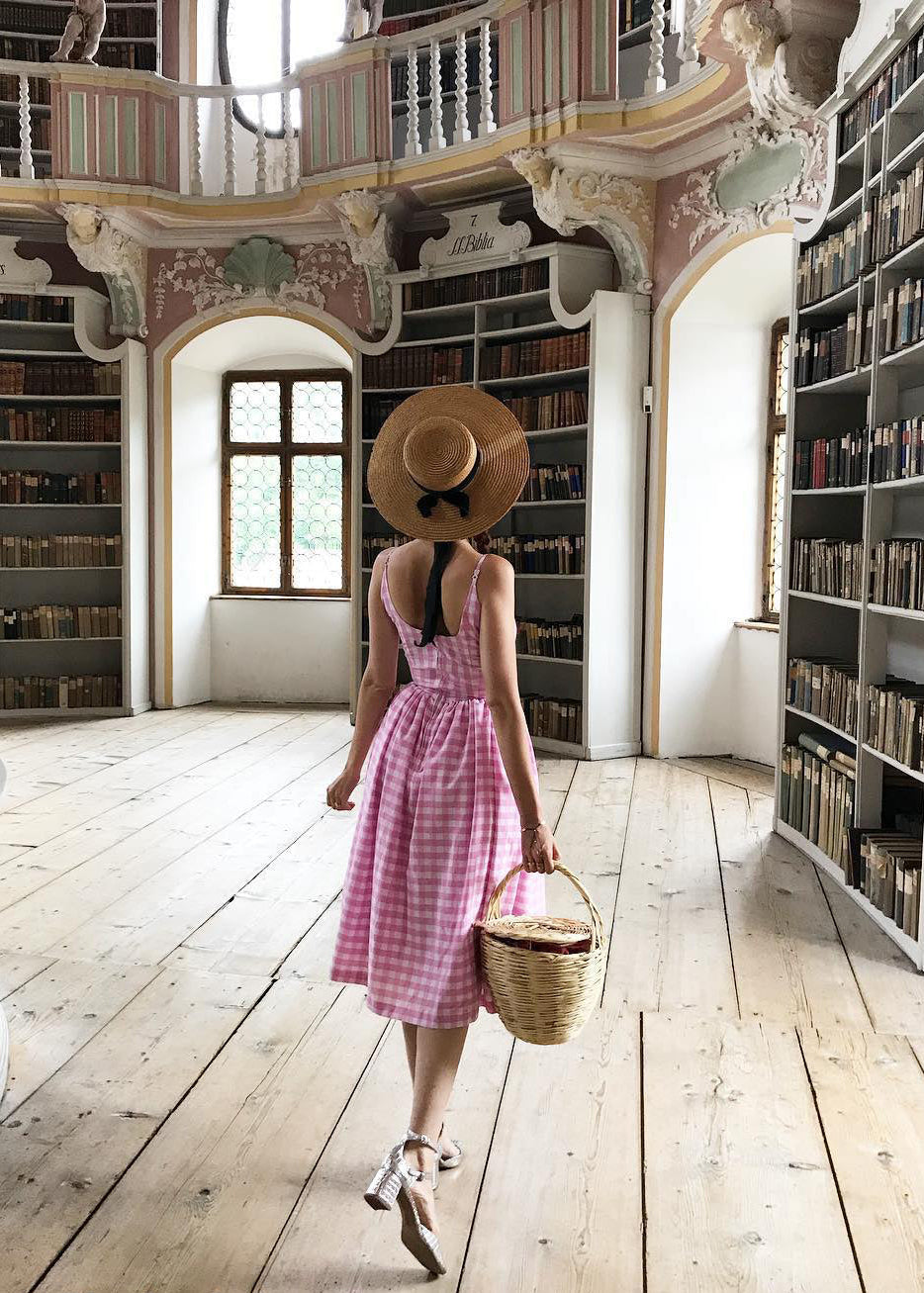 Woman in a pink dress and straw hat walking through a large, ornate library.