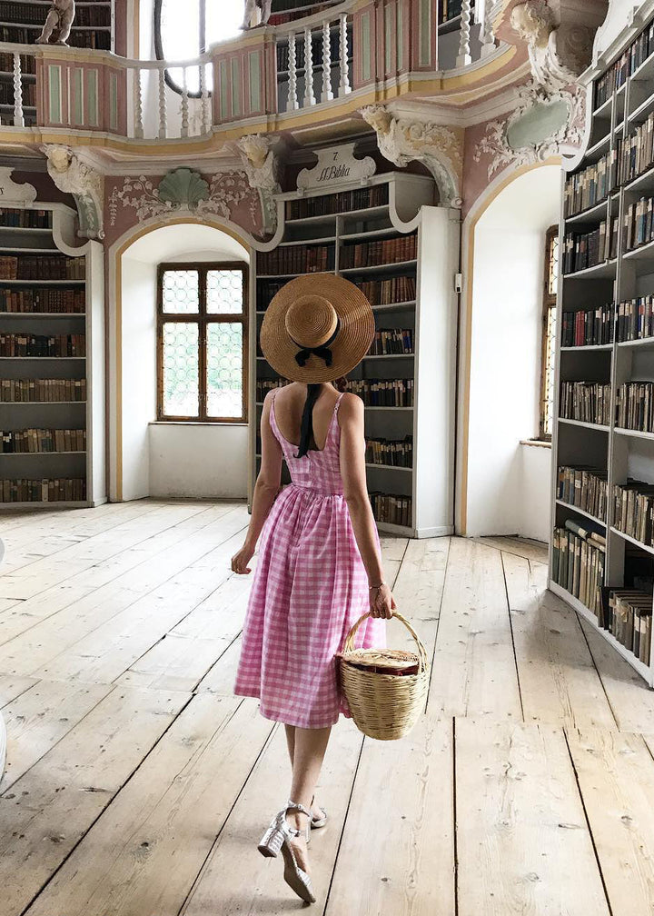 Woman in a pink dress and straw hat walking through a large, ornate library.
