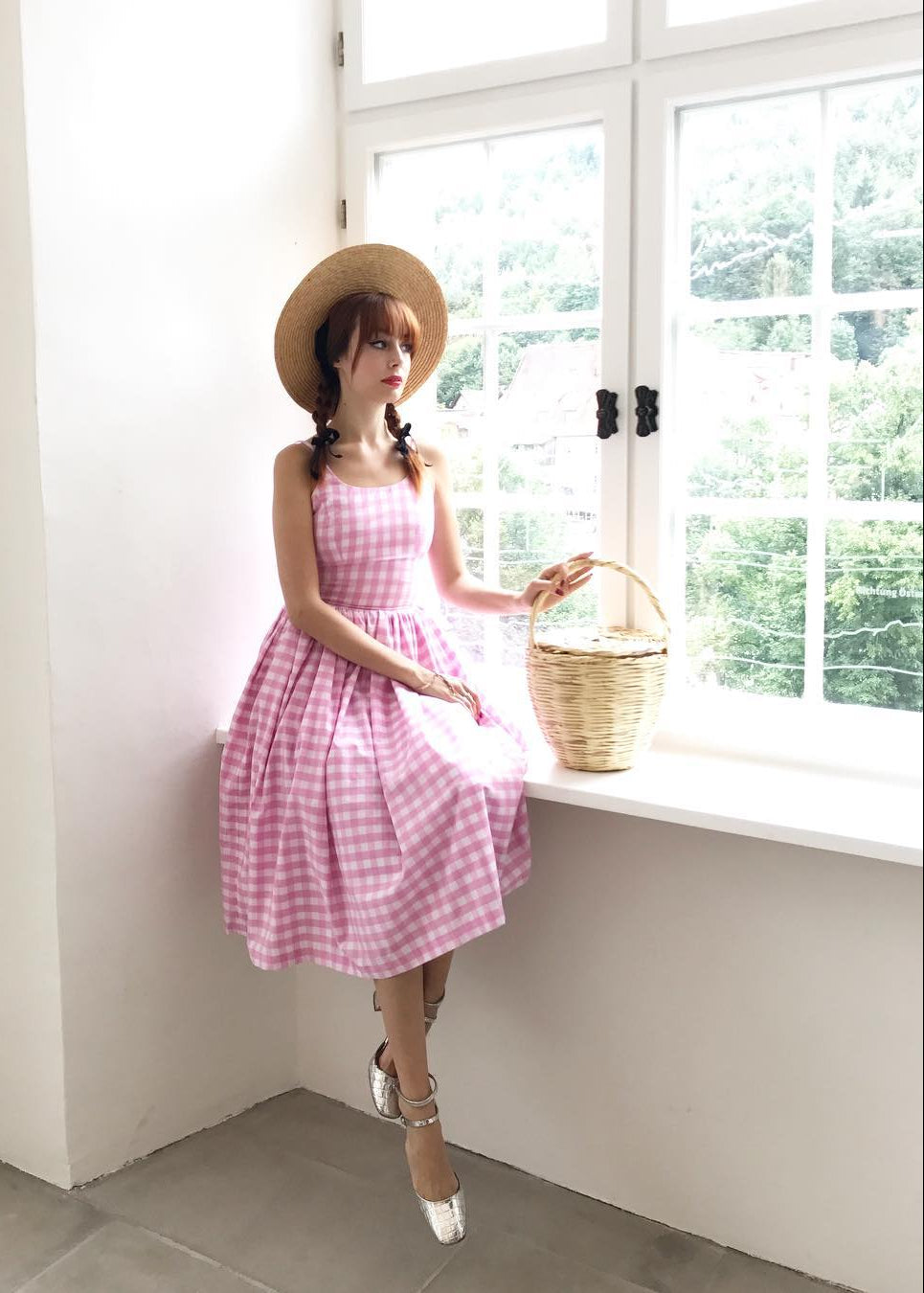 Woman in a pink checkered dress and straw hat sitting by a window holding a basket.