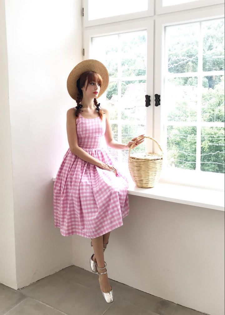 Woman in a pink checkered dress and straw hat sitting by a window holding a basket.