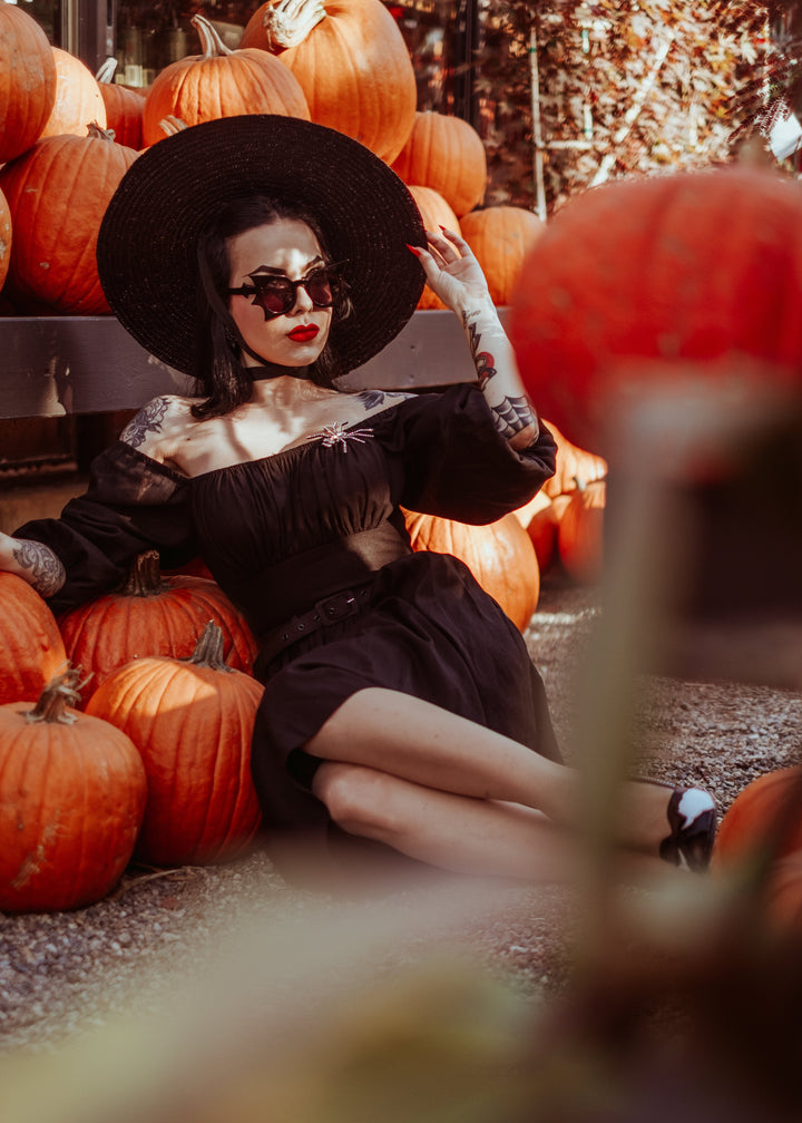 A woman in a black witch-style outfit with a wide-brimmed black hat, black sunglasses, and red lipstick sits among orange pumpkins outdoors.