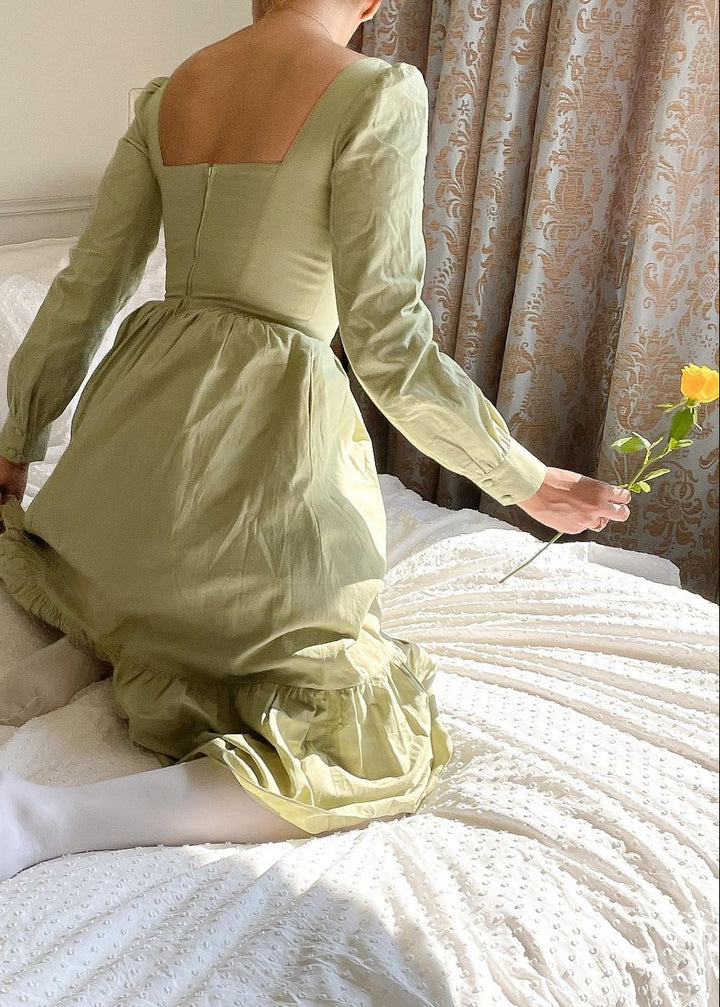 Person wearing a light green dress sitting on a bed holding a yellow flower.