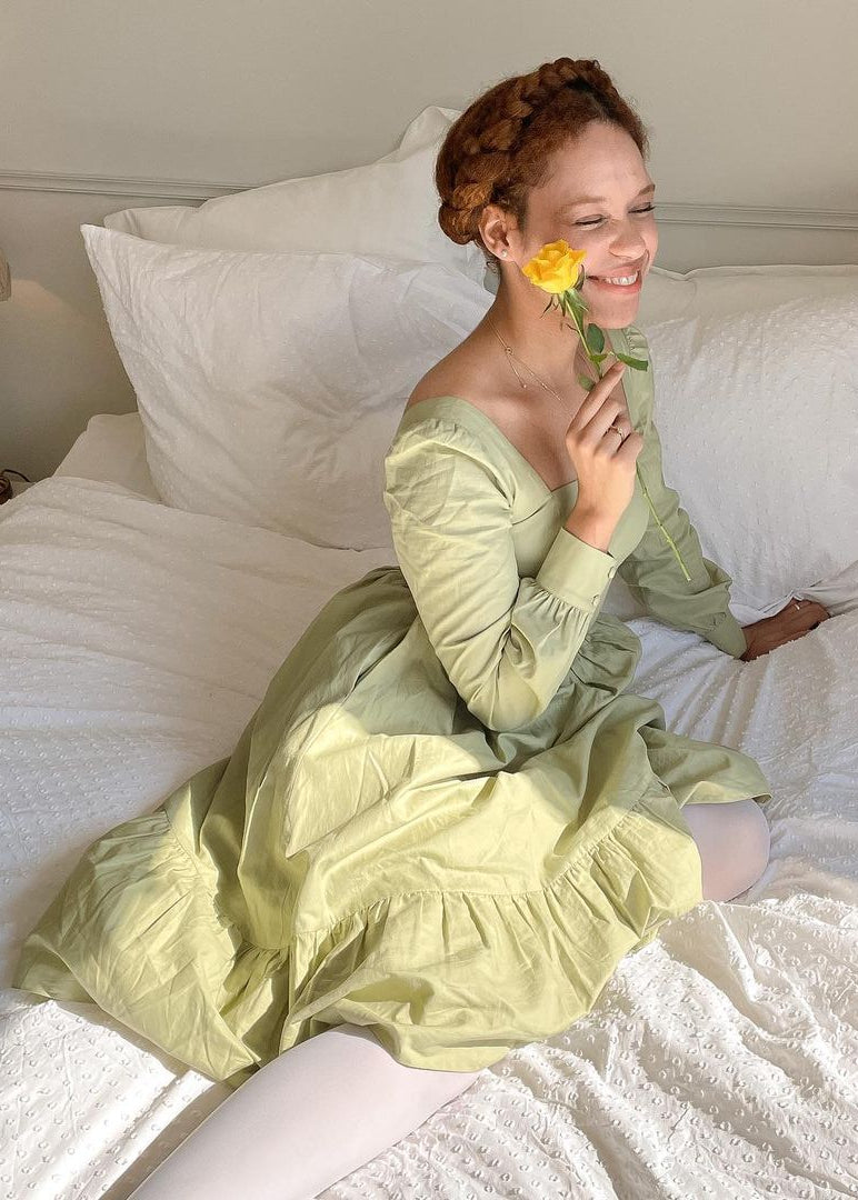 Woman in a light green dress holding a yellow flower on a bed.