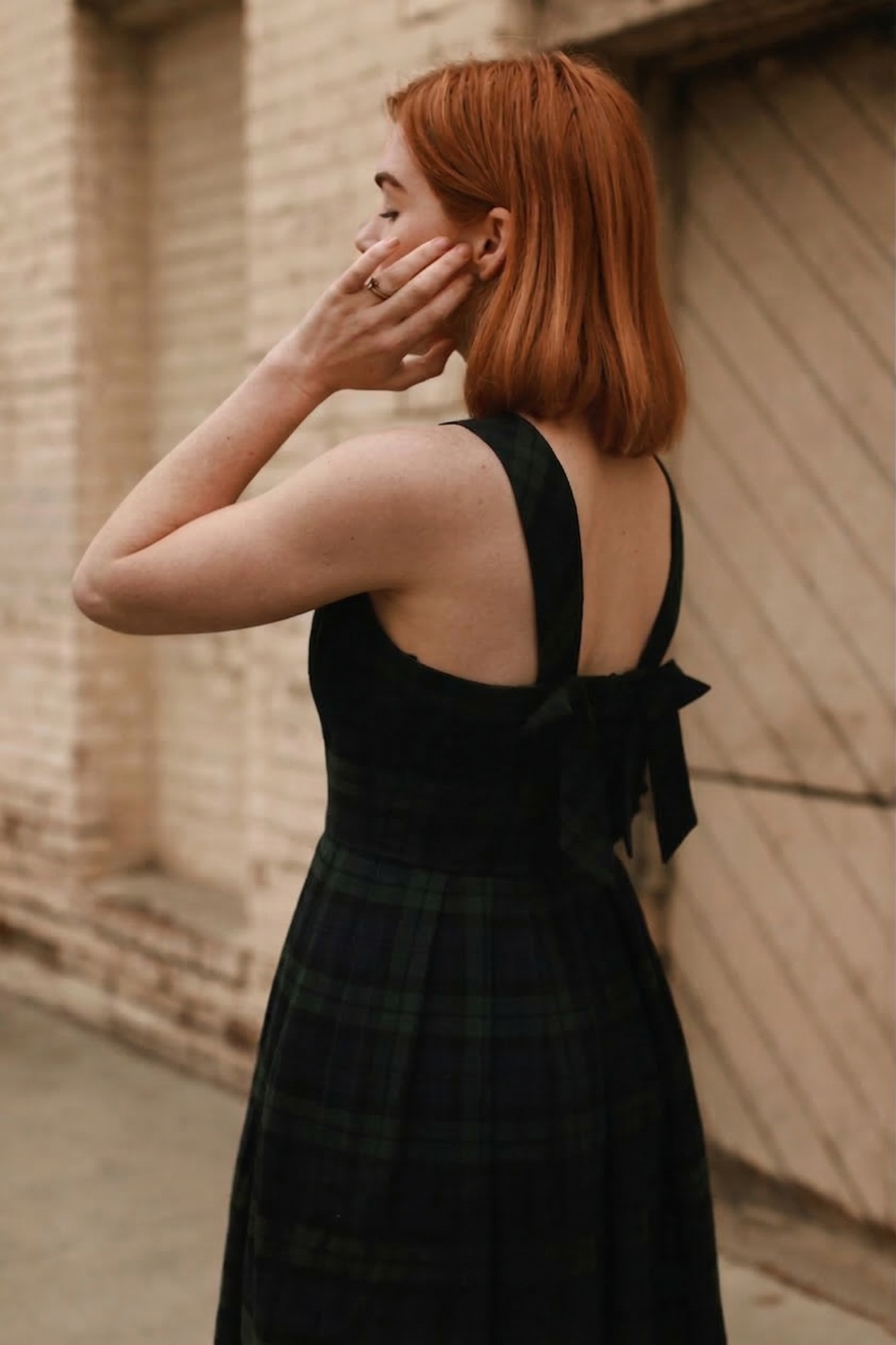 Woman in a plaid dress with a bow standing against a textured wall.