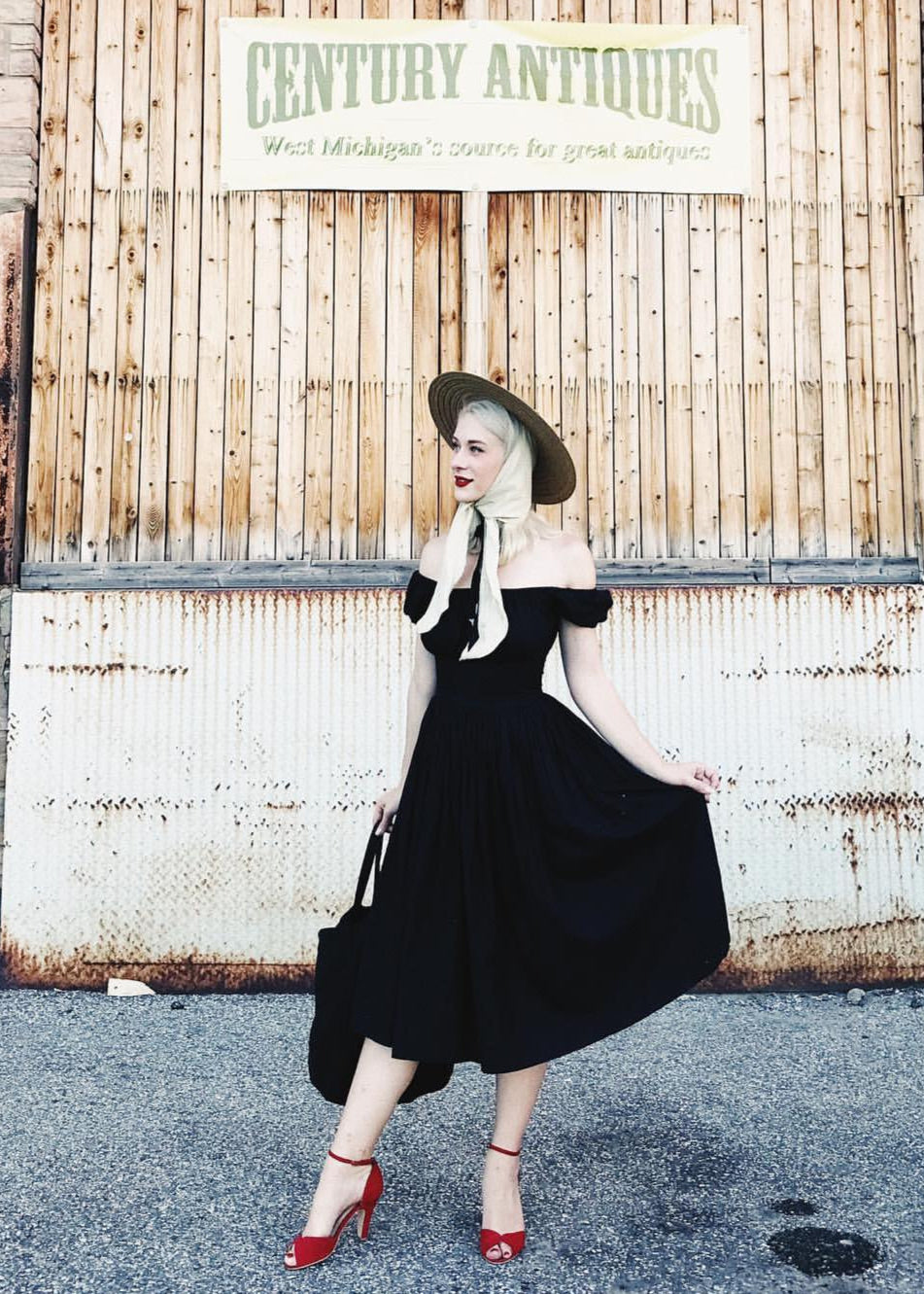 Woman in a black dress and red shoes standing in front of a rustic wooden wall with 'Century Antiques' sign.