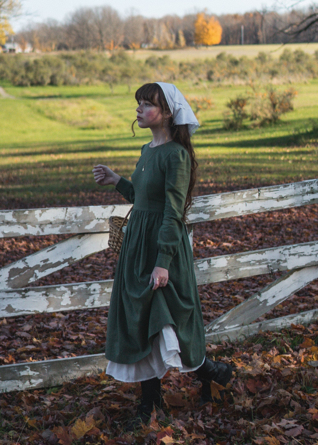 Person in a green dress standing in a field with a wooden fence and trees in the background