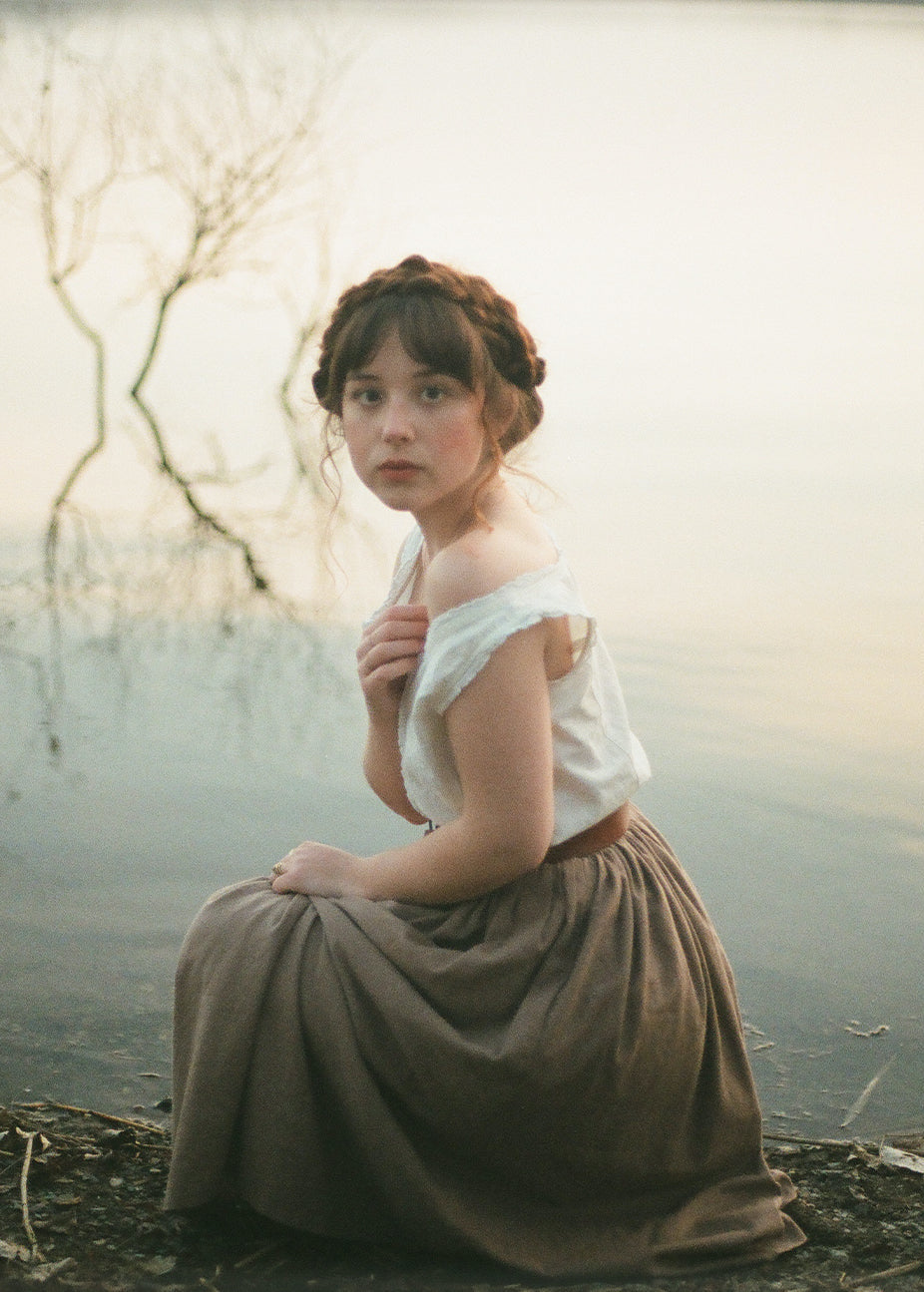 Woman in a white top and brown skirt sitting by a body of water with bare trees in the background.