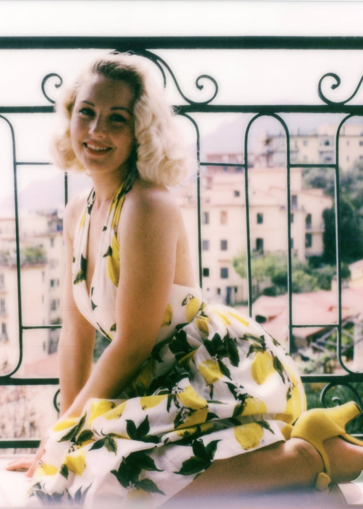 Woman in a yellow and white dress sitting on a balcony with a cityscape in the background