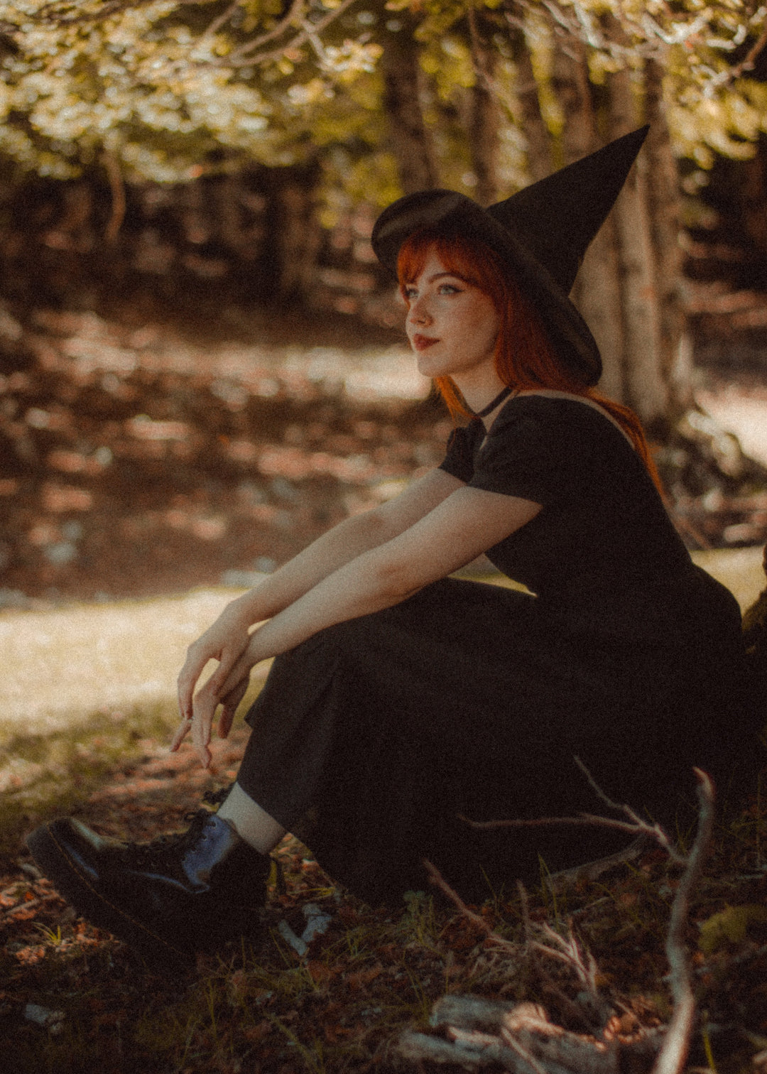 A woman with red hair wearing a black witch hat and a black dress sits on the ground in a sun-dappled forest setting.