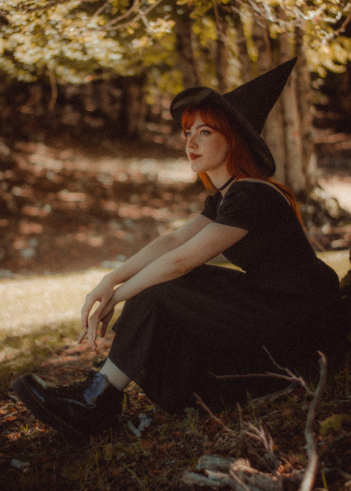 A woman with red hair wearing a black witch hat and a black dress sits on the ground in a sun-dappled forest setting.