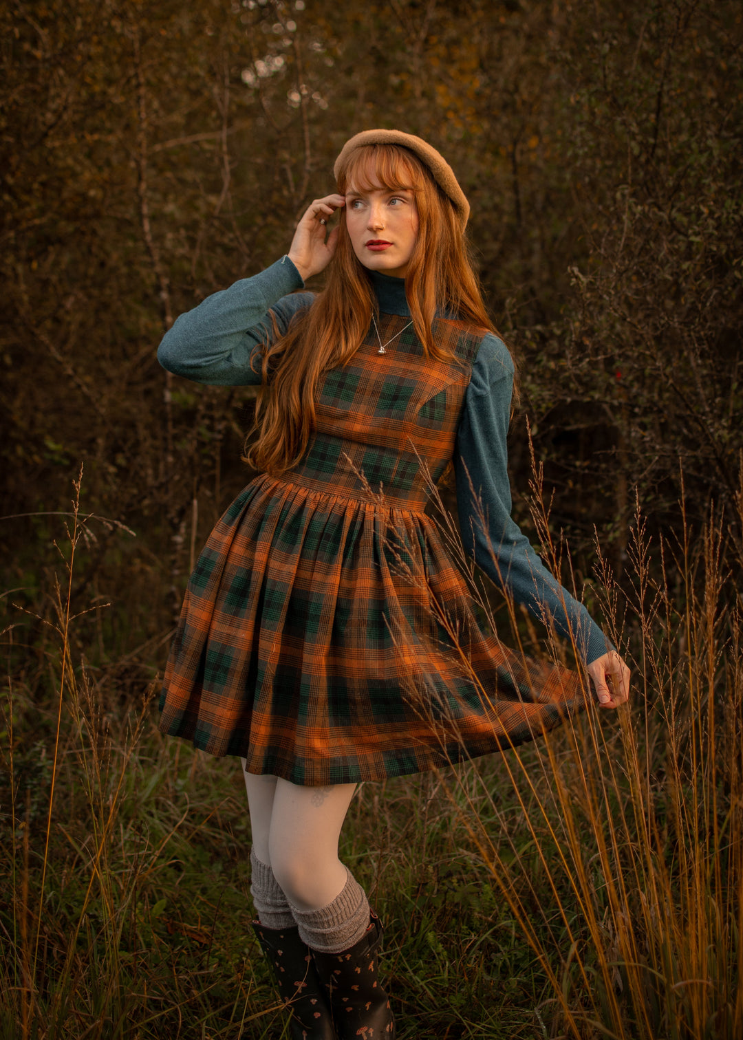 Woman in a plaid dress and beret standing in tall grass with a dark background